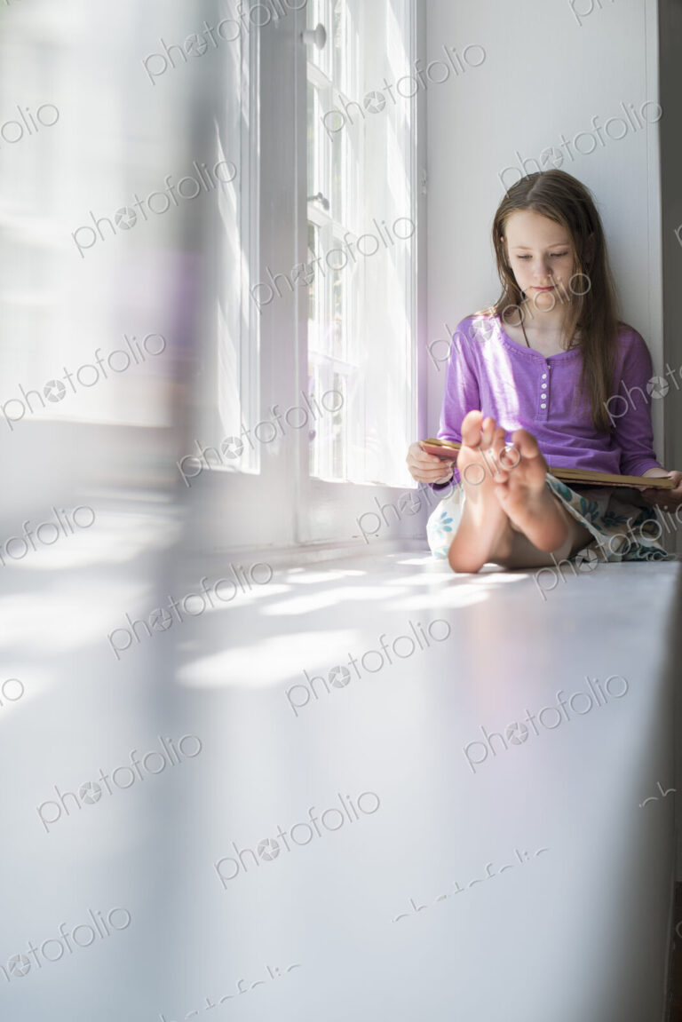 Girl sitting by a window, reading a book.