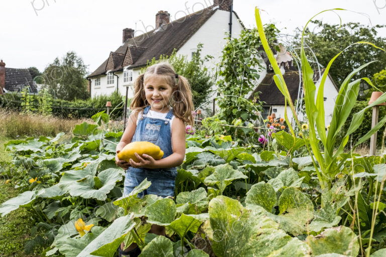 Girl standing in a vegetable patch in a garden, holding yellow gourd, smiling at camera.