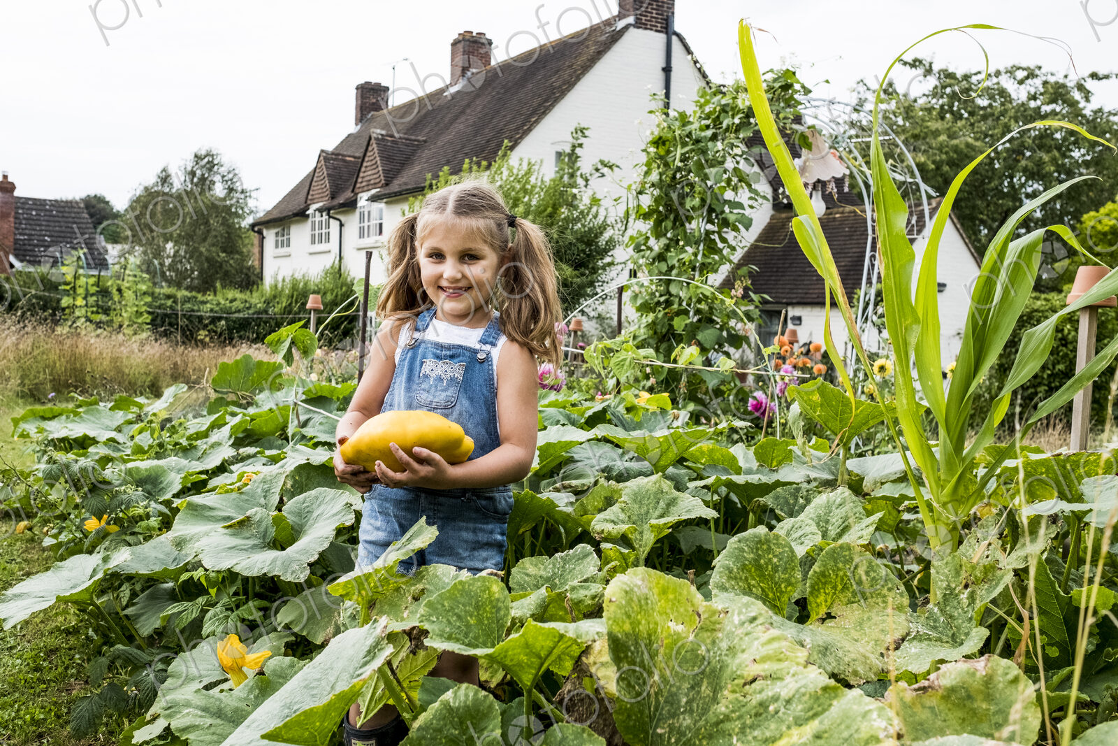Girl standing in a vegetable patch in a garden, holding yellow gourd, smiling at camera.