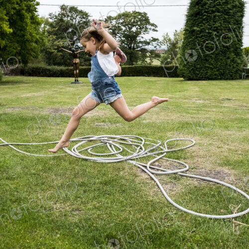 Girl wearing denim dungarees jumping over a garden hose on a lawn.