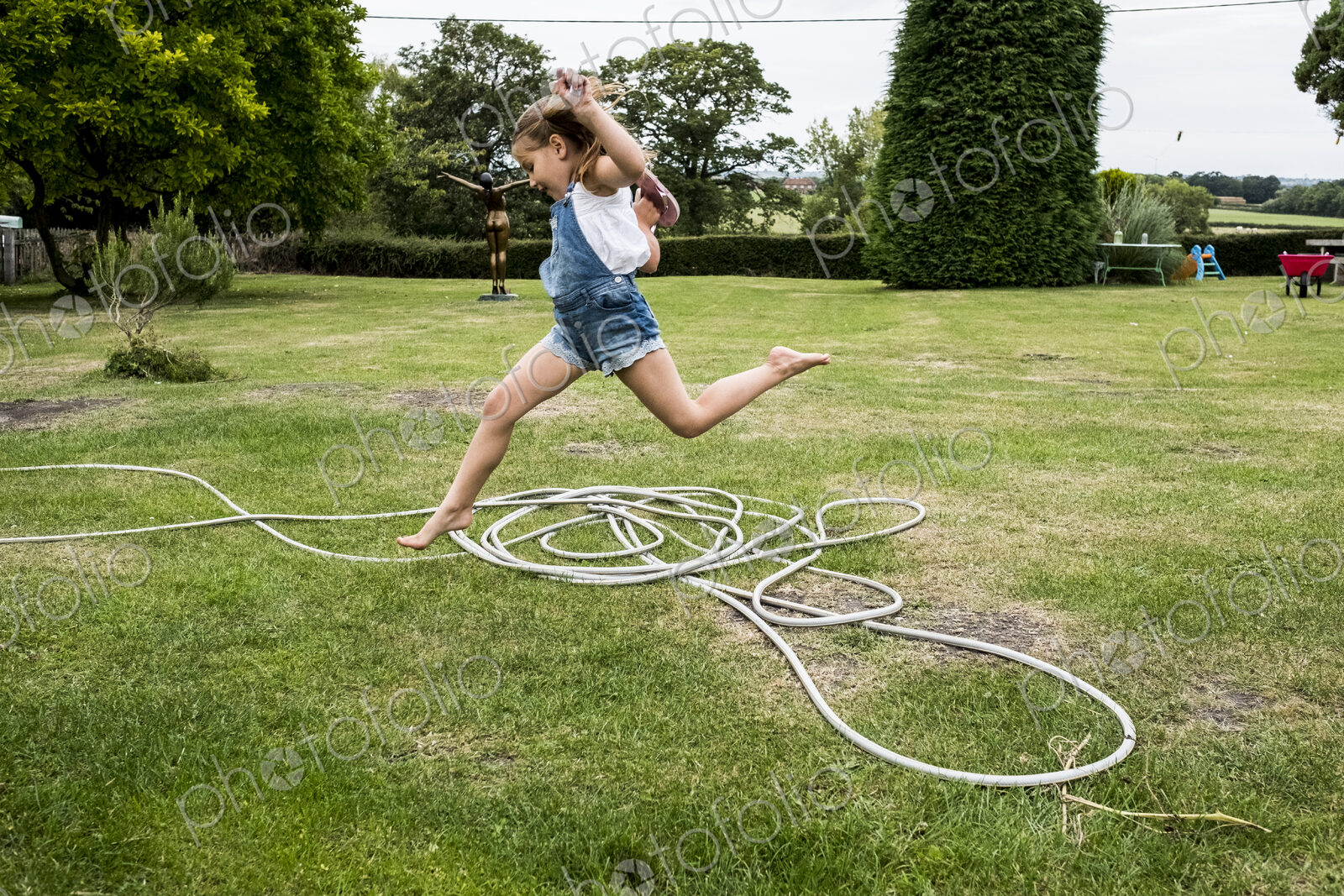 Girl wearing denim dungarees jumping over a garden hose on a lawn.