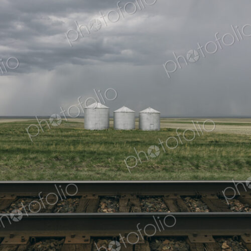 Grain silos and storm clouds over vast areas of farmland and prairie, train tracks in…