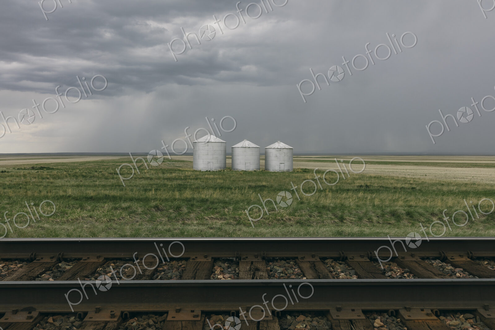 Grain silos and storm clouds over vast areas of farmland and prairie, train tracks in…