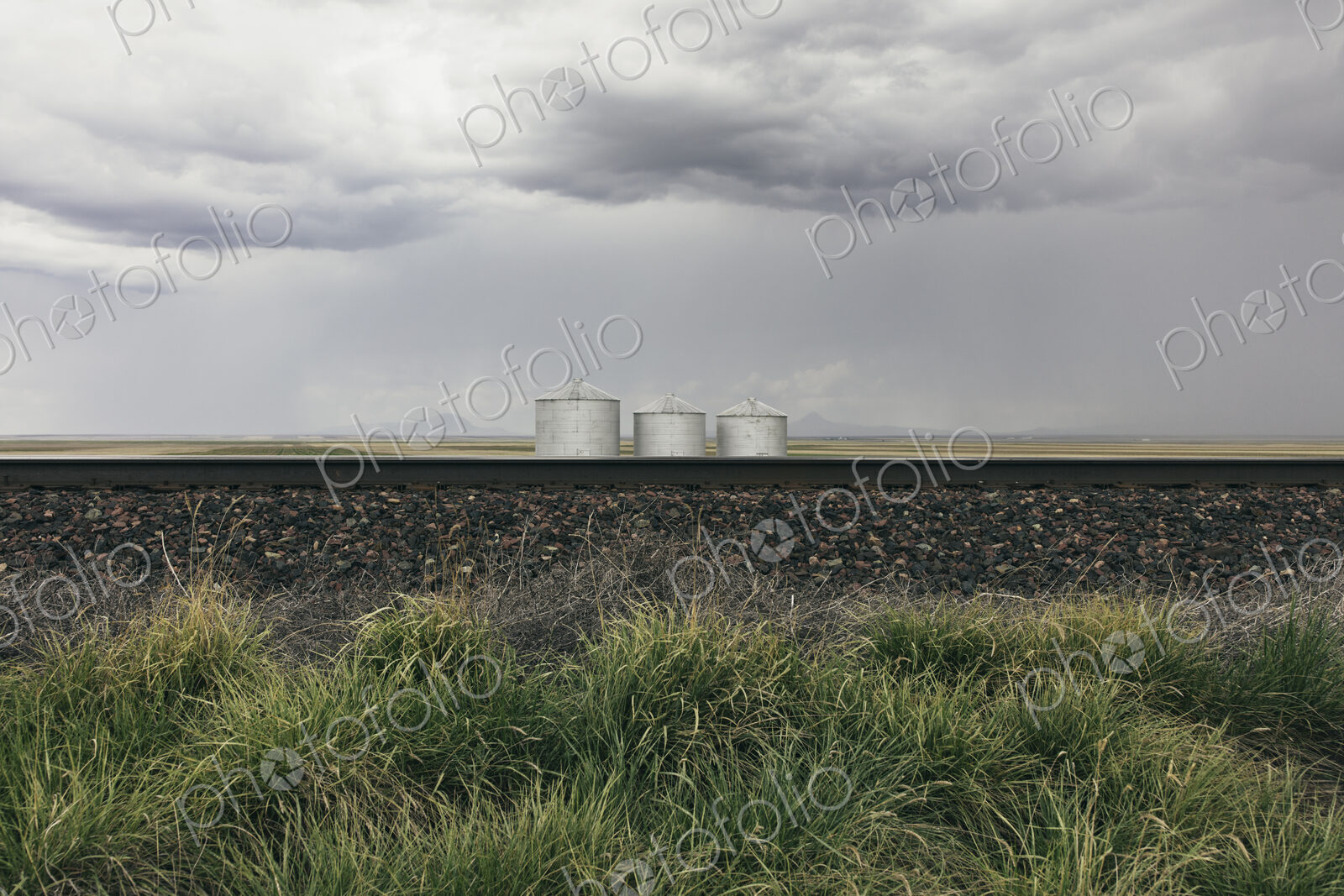 Grain silos and storm clouds over vast farmland and prairie, train tracks in foreground