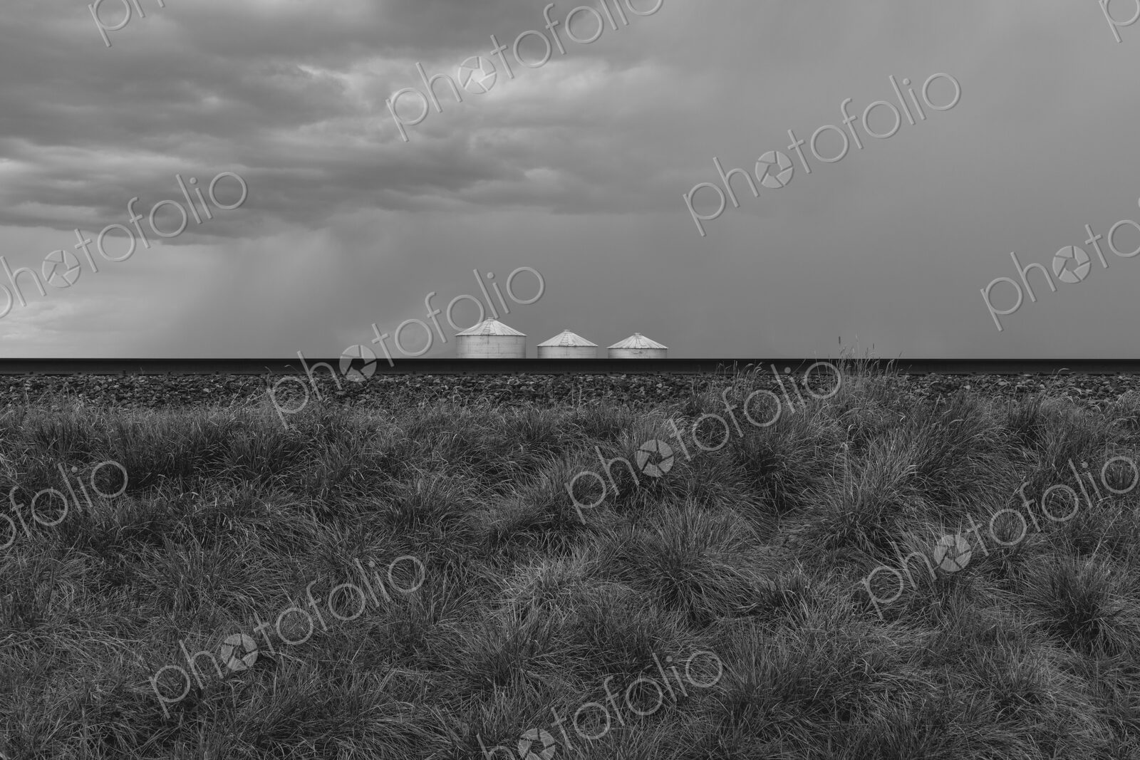 Grain silos and storm clouds over vast farmland and prairie, train tracks in foreground