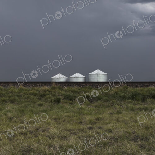 Grain silos and storm clouds over vast farmland and prairie, train tracks in foreground