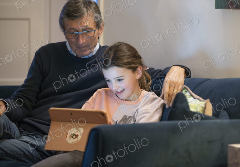 Grandfather and granddaughter sitting together on sofa looking at tablet.