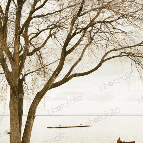 Grandmother and toddler granddaughter sitting on a park bench, facing the lake at… Grandmother and toddler granddaughter sitting on a park bench, facing the lake at…