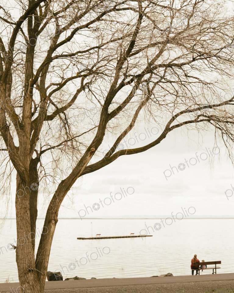 Grandmother and toddler granddaughter sitting on a park bench, facing the lake at…