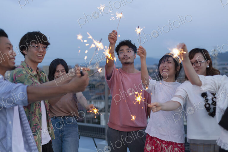 Group of young Japanese men and women with sparklers on a rooftop in an urban setting.