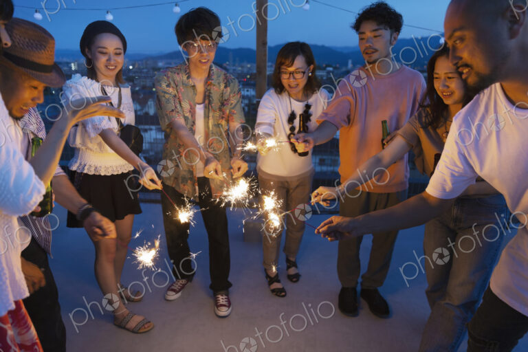 Group of young Japanese men and women with sparklers on a rooftop in an urban setting.