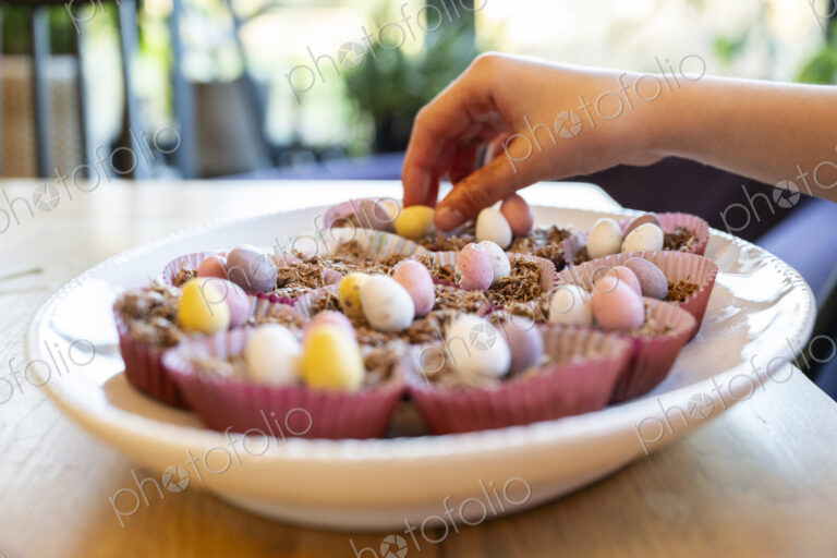 Hand putting decorations on to Easter cup cakes in dish on kitchen table.