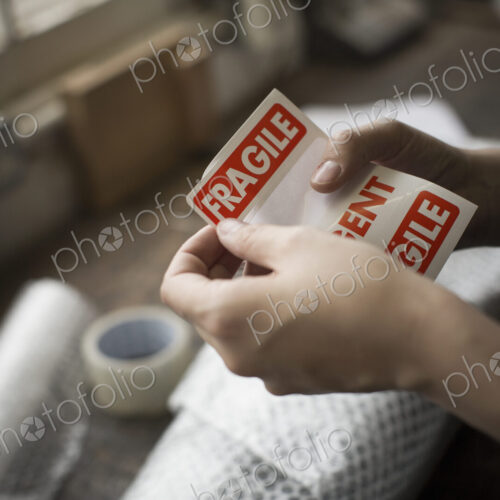 Hands holding a red Fragile sticker to stick it on a brown paper package on a work bench. Hands holding a red Fragile sticker to stick it on a brown paper package on a work bench.