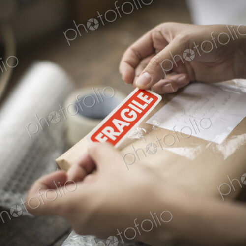 Hands holding a red Fragile sticker to stick it on a brown paper package on a work bench. Hands holding a red Fragile sticker to stick it on a brown paper package on a work bench.