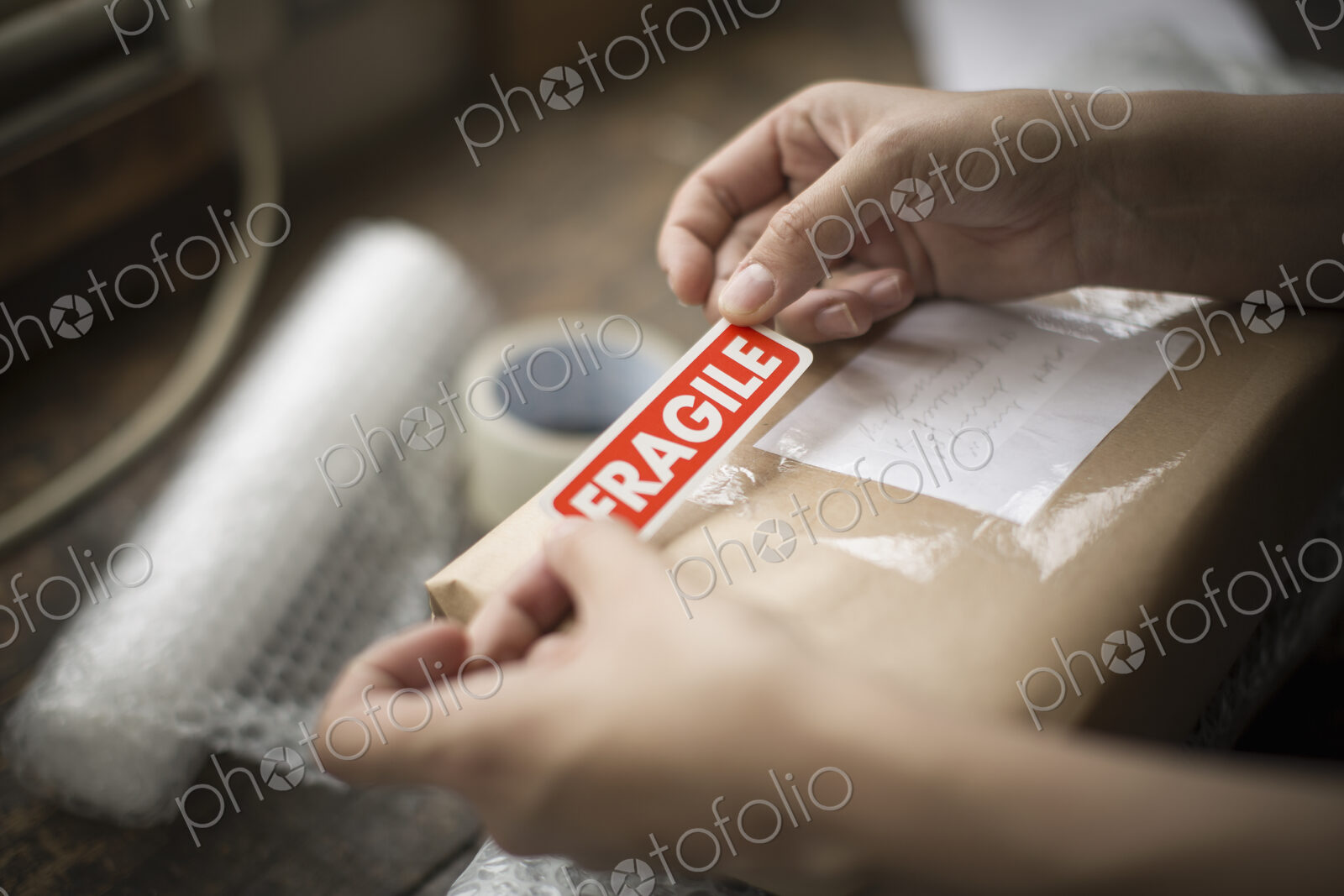Hands holding a red Fragile sticker to stick it on a brown paper package on a work bench.