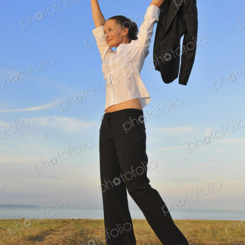 Happy businesswoman with arms raised holding suit jacket walking on sandy beach, freedom.