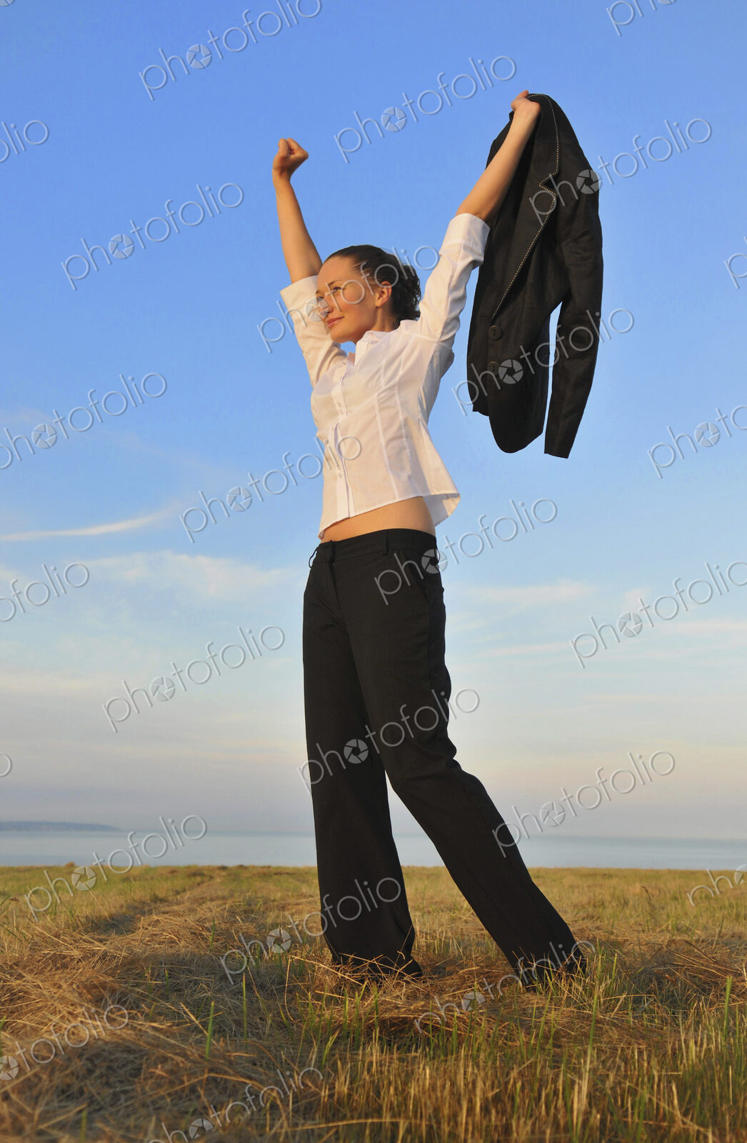 Happy businesswoman with arms raised holding suit jacket walking on sandy beach, freedom.