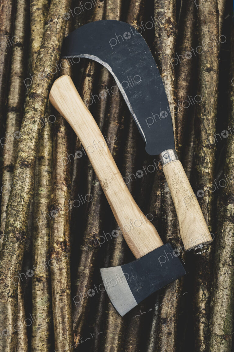 High angle close up of axe and bill hook lying on bunch of wooden stakes used in…