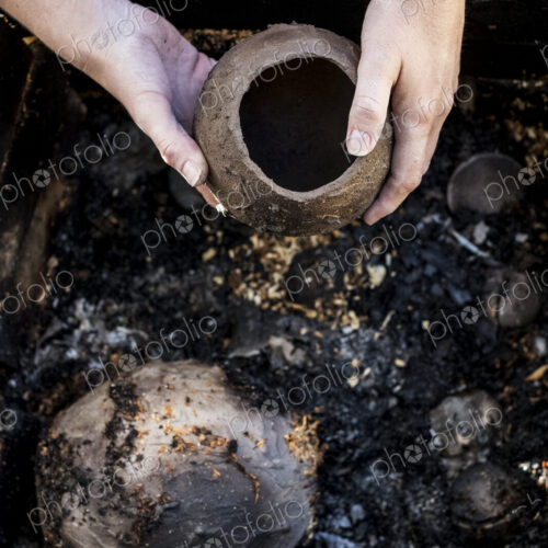 High angle close up of ceramic artist sitting next to outdoor smoke fire pit, working…