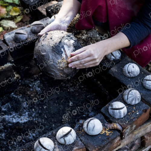 High angle close up of ceramic artist sitting next to outdoor smoke fire pit, working…