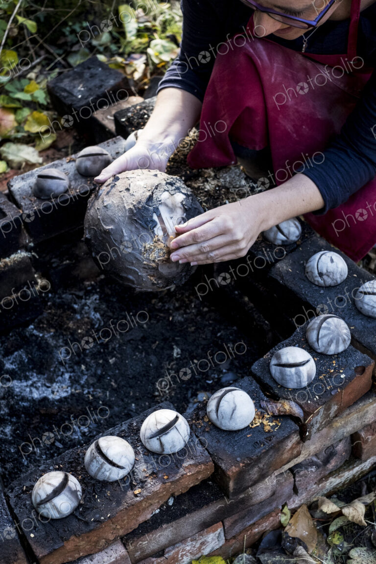 High angle close up of ceramic artist sitting next to outdoor smoke fire pit, working…