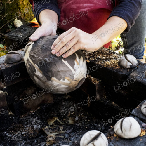 High angle close up of ceramic artist sitting next to outdoor smoke fire pit, working…