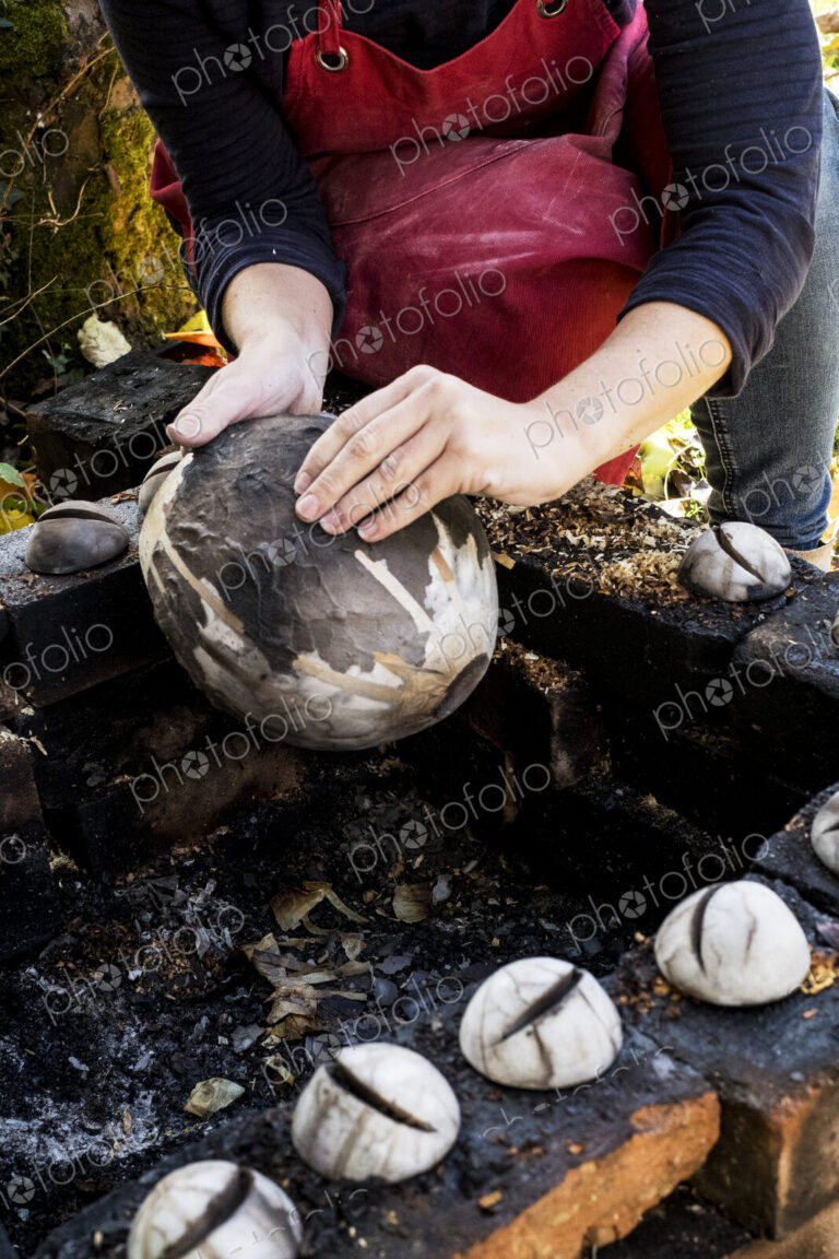 High angle close up of ceramic artist sitting next to outdoor smoke fire pit, working…
