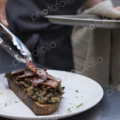 High angle close up of chef preparing toasted bread with bacon and mushrooms in an… High angle close up of chef preparing toasted bread with bacon and mushrooms in an…