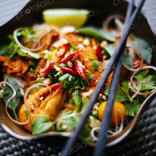High angle close up of chopsticks on bowl with Asian food containing noodles, prawns,… High angle close up of chopsticks on bowl with Asian food containing noodles, prawns,…