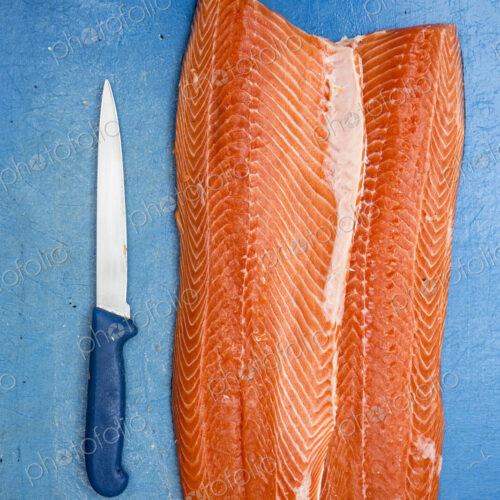 High angle close up of fillets of fresh salmon and knife on blue chopping board.