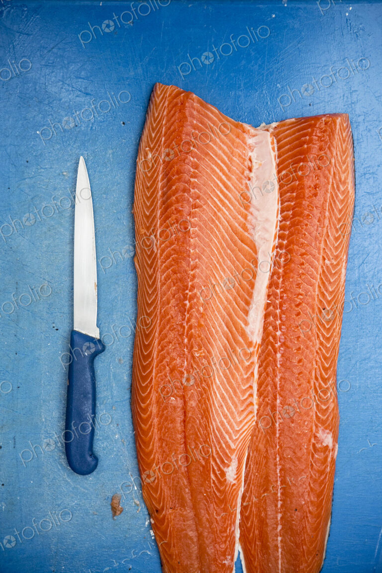 High angle close up of fillets of fresh salmon and knife on blue chopping board.