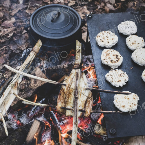 High angle close up of fish on skewers and freshly made buns over a campfire.