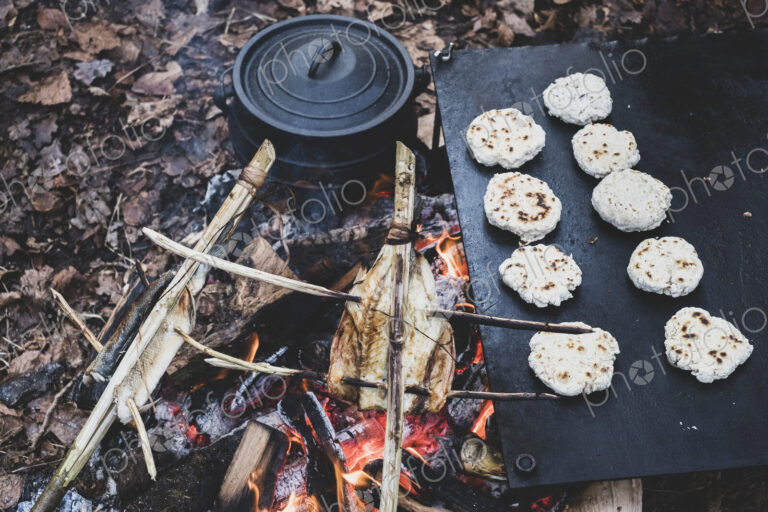 High angle close up of fish on skewers and freshly made buns over a campfire.