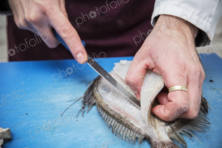 High angle close up of fishmonger filleting John Dory fish with knife on blue chopping…