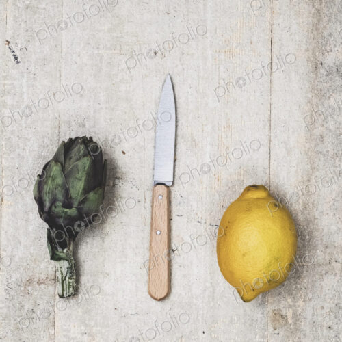 High angle close up of fresh lemon, artichoke and kitchen knife on grey background.