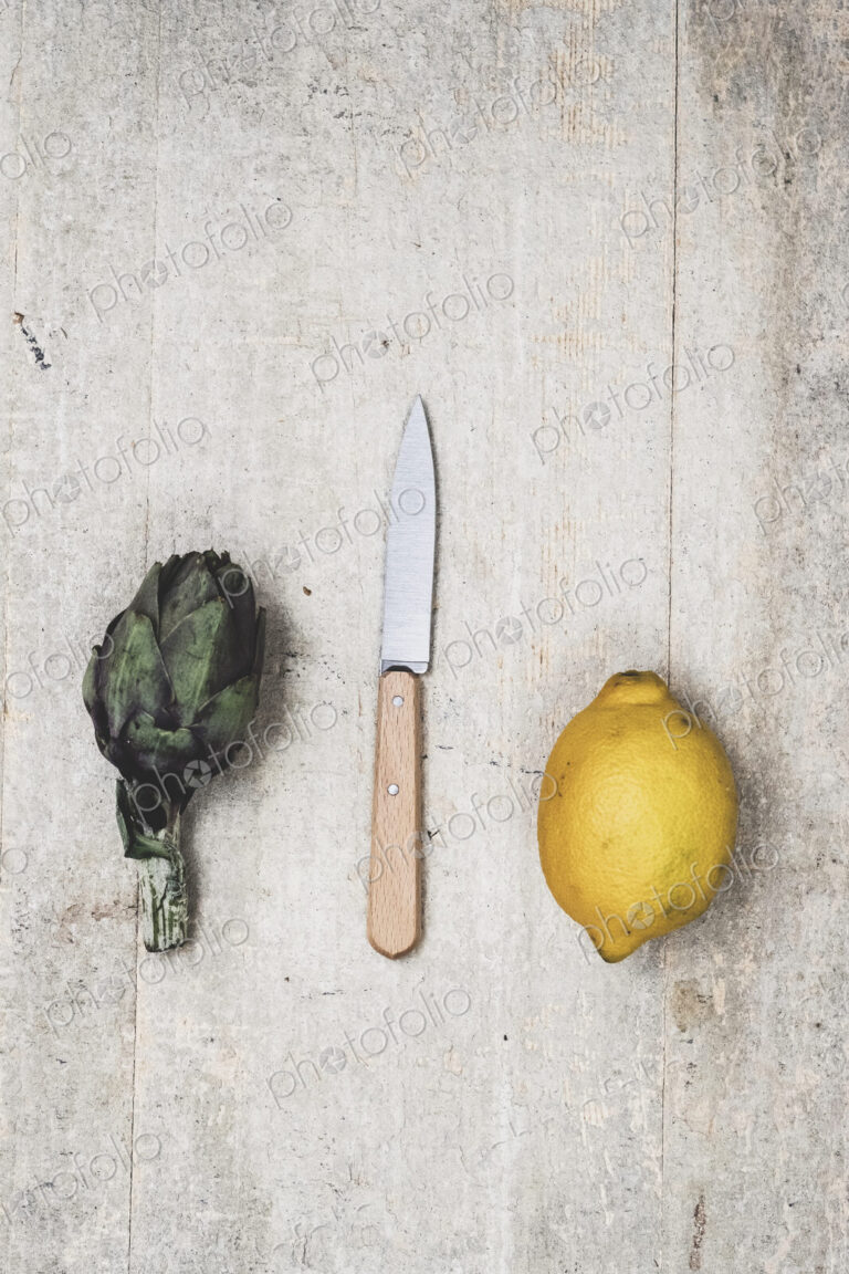 High angle close up of fresh lemon, artichoke and kitchen knife on grey background.