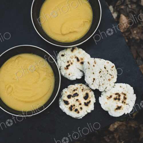 High angle close up of freshly made bread buns and bowls of soup over a campfire.