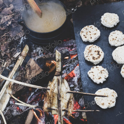 High angle close up of freshly made buns and fish on skewers over a campfire, and a…