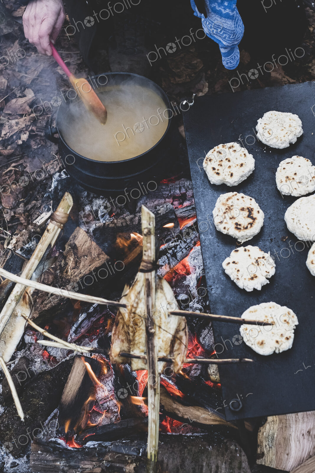 High angle close up of freshly made buns and fish on skewers over a campfire, and a…