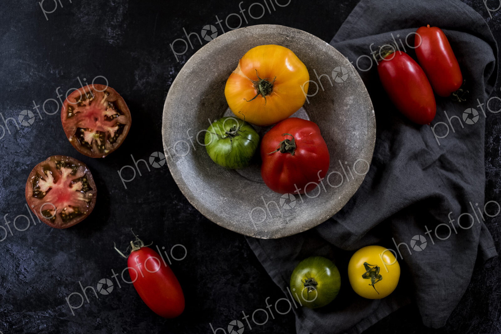 High angle close up of grey plate and cloth and a selection of fresh tomatoes on black…