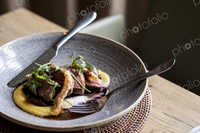 High angle close up of grey plate of meat with mashed potato, crackling and rocket…
