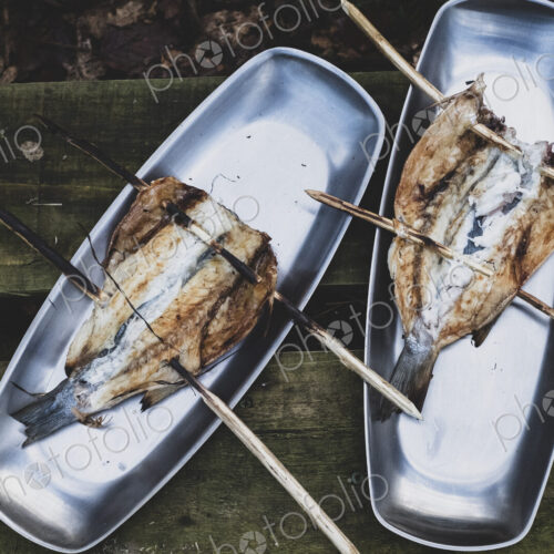 High angle close up of grilled fish on wooden skewers on metal plates, cooking outdoors