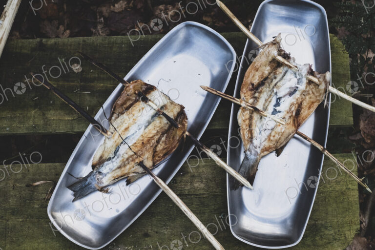 High angle close up of grilled fish on wooden skewers on metal plates, cooking outdoors