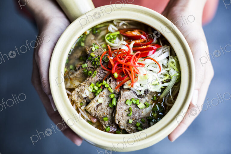 High angle close up of hands holding bowl with Asian soup containing rice vermicelli,…