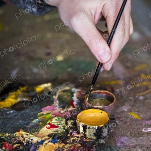 High angle close up of human hand dipping paintbrush into small pot of yellow oil paint. High angle close up of human hand dipping paintbrush into small pot of yellow oil paint.