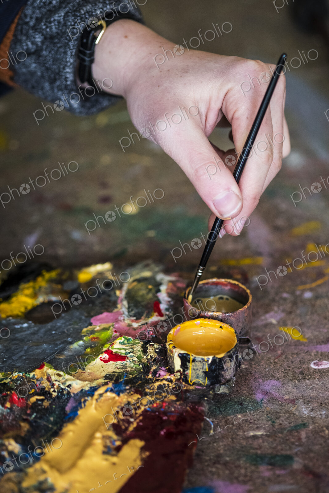 High angle close up of human hand dipping paintbrush into small pot of yellow oil paint.