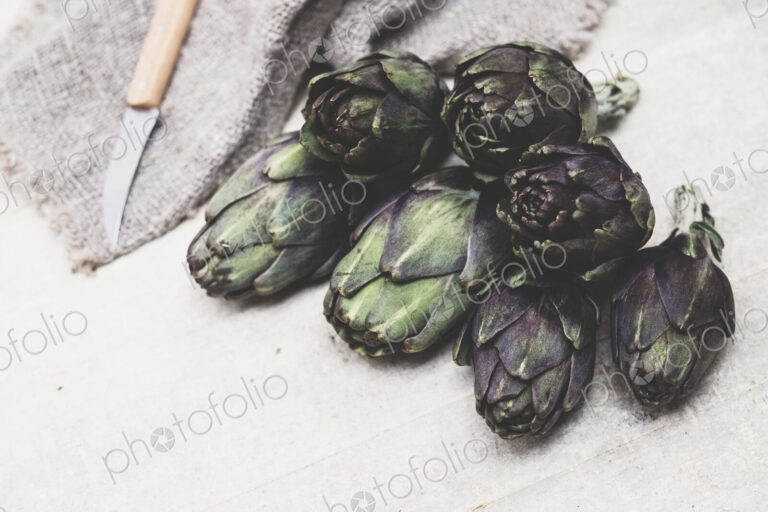 High angle close up of kitchen knife and fresh artichokes on grey background.