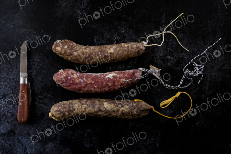 High angle close up of knife and selection of three salamis on black background.