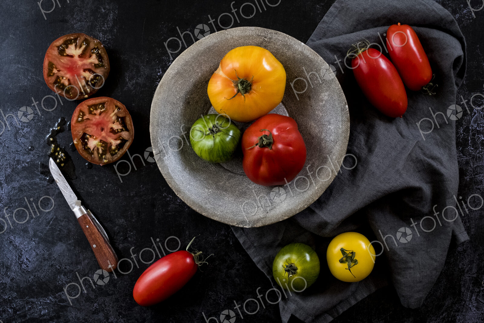 High angle close up of knife, grey plate and cloth and a selection of fresh tomatoes…