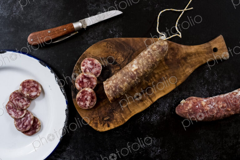 High angle close up of knife, sliced salami on wooden cutting board and white enamel…